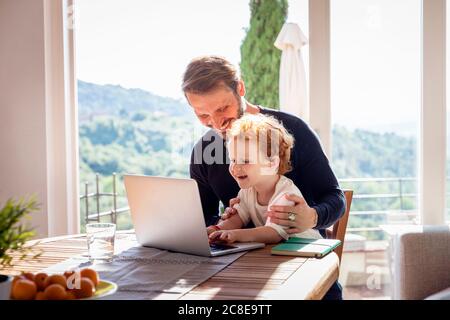 Lächelnder Mann, der seinen Sohn mit einem Laptop ansieht, während er sich dagegen ansieht Fenster im Wohnzimmer Stockfoto