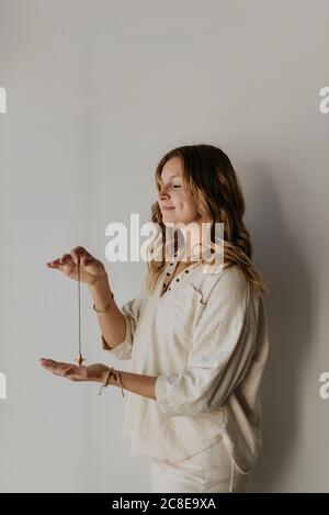 Frau mit mittlerem Erwachsenen, die das Pendel hält, meditiert, während sie gegen Weiß steht Wand Stockfoto