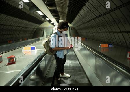 Mann mit Schutzmaske, der auf der Rolltreppe steht und auf das Mobiltelefon schaut, London, Großbritannien Stockfoto