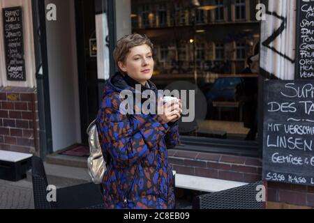 Porträt einer träumenden Frau mit Rucksack und Kaffee zum mitnehmen Vor einem Café Stockfoto