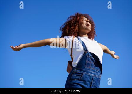 Sorglose Afro-Frau mit ausgestreckten Armen, die gegen klares Blau steht Himmel Stockfoto