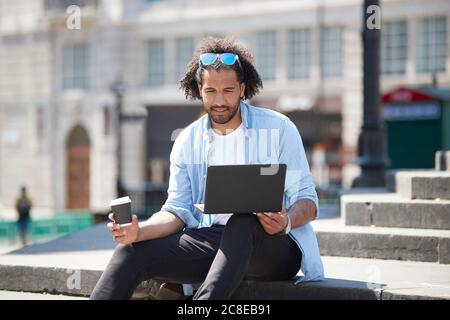 Porträt eines jungen Mannes mit Kaffee zu gehen sitzen auf einer Treppe im Freien Blick auf Laptop, London, Großbritannien Stockfoto