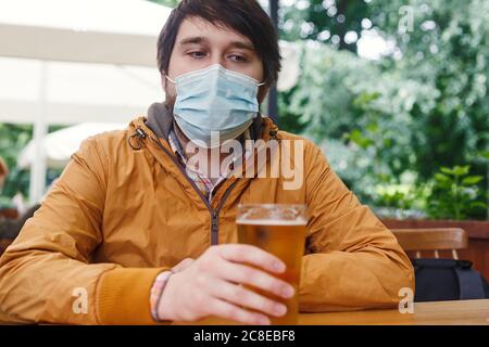 Kaukasischer Mann mit einem Glas Bier in einer Arznei-Maske auf der Veranda des Cafés sitzen. Neues normales Konzept Stockfoto