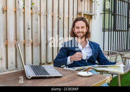 Lächelnd hübscher junger Geschäftsmann, der Zeitung liest, während er die Kaffeetasse hält Mit dem Laptop am Tisch im Café Stockfoto