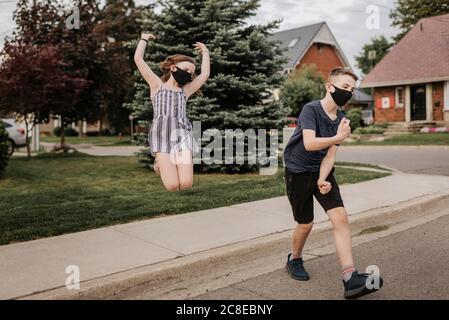 Unbeschwerte Geschwister tragen Masken, tanzen und springen auf der Straße hinein Stadt Stockfoto