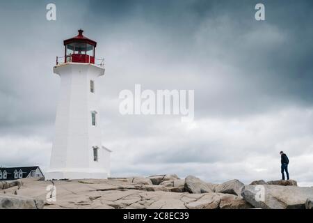 Mann, der in der Nähe von Peggys Point Lighthouse auf Felsformation gegen bewölkten Himmel steht, Nova Scotia, Kanada Stockfoto