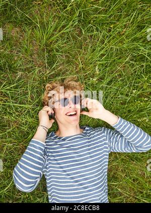 Porträt eines lachenden jungen Mannes mit lockigen blonden Haaren liegend Auf einer Wiese Musik hören mit Kopfhörern