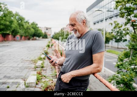 Lächelnder älterer Mann, der Smartphone benutzt, während er am Geländer steht In der Stadt Stockfoto