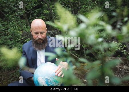 Kahlbärtiger Geschäftsmann hält Globus, während er im Wald sitzt Stockfoto