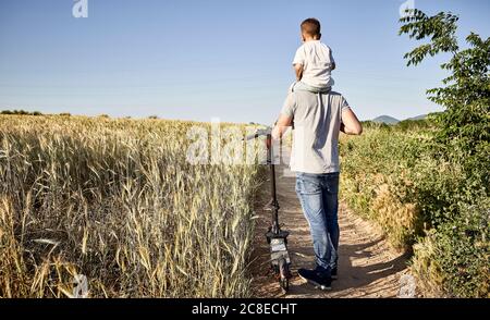 Vater trägt Sohn auf den Schultern beim Gehen mit Roller schieben Inmitten von Pflanzen Stockfoto