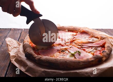 Eine Hand schneidet Pizza mit rundem Messer auf einem dunklen Holztisch Stockfoto