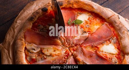 Eine Hand schneidet Pizza mit rundem Messer auf einem dunklen Holztisch Stockfoto