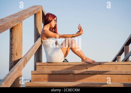 Teenager-Mädchen, die Selfie durch Smartphone während des Sitzens auf Holztreppen gegen klaren Himmel Stockfoto