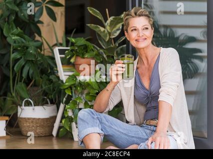 Porträt einer entspannten reifen Frau, die an der offenen Terrassentür sitzt Mit Glas grünem Smoothie Stockfoto