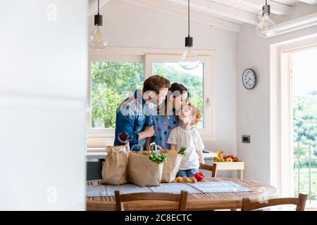 Lächelnder Mann, der Frau beim Küssen des Sohnes beim Stehen ansieht Esstisch mit Lebensmitteln in der Küche Stockfoto