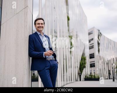 Lächelnder reifer Geschäftsmann mit Smartphone lehnt sich an ein Gebäude in Die Stadt Stockfoto