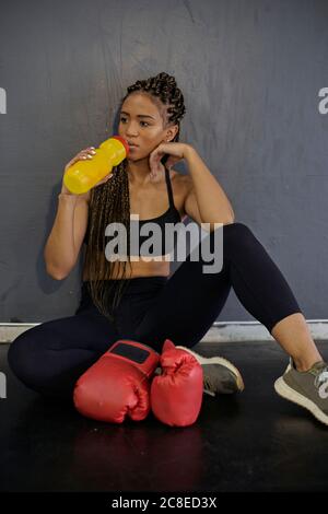 Weibliche Athletin mit geflochtenen Haaren Trinkwasser beim Sitzen gegen Wand im Fitnessraum Stockfoto