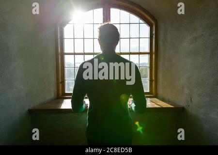 Rückansicht eines jungen Mannes, der durch ein geschlossenes Vintage-Holzfenster mit einströmendem Sonnenlicht schaute Stockfoto