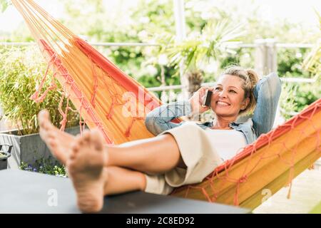 Portrait von glücklichen reifen Frau auf dem Telefon entspannen in Hängematte auf der Terrasse Stockfoto