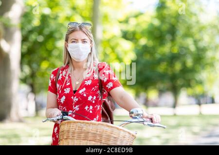 Frau trägt Maske, während sie mit dem Fahrrad im öffentlichen Park steht Stockfoto