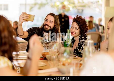Lächelnder junger bärtiger Mann, der Selfie mit einer sitzenden Frau nimmt Tisch im Restaurant Stockfoto