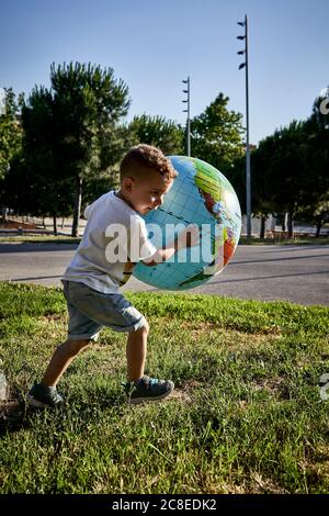 Sorgloser Junge hält Globus während auf grasbewachsenem Land laufen während Sonniger Tag Stockfoto