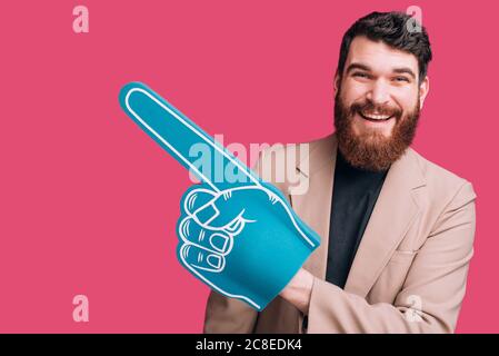 Schöner bärtiger Mann trägt Jacke und einen blauen Fan-Schaum-Handschuh auf rosa Hintergrund. Stockfoto