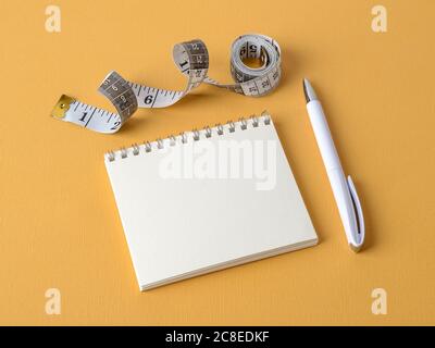 Öffnen Sie den Papiernotizblock mit Kopierplatz, weißem Stift und einer Rolle Maßband mit Zentimetern und Zoll auf einem gelben strukturierten Hintergrund. Speicherplatz kopieren. Stockfoto