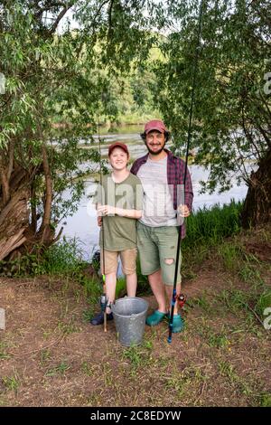 Glücklicher Vater und Sohn halten Angelruten während des Stehens an Flussufer Stockfoto