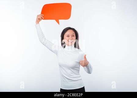 Lächelnde junge Frau hält eine orangefarbene Blasenrede über ihrem Kopf auf weißem Hintergrund. Stockfoto