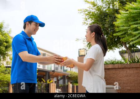 Asiatische Lieferung junger Mann in blauen Uniform Lächeln und hält eine Pappkartons vor Haus und asiatische Frau Annahme einer Lieferung von Boxen aus liefern Stockfoto