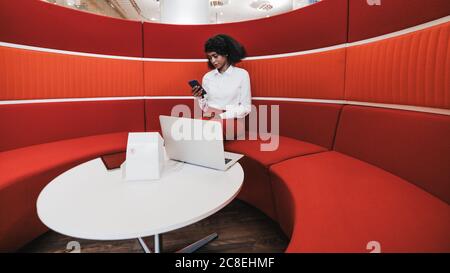 Die schöne afroamerikanische Unternehmerin sitzt in einem gemütlichen Büro Coworking Area und unterhält Geschäftsgespräche über ihr Smartphone und den Laptop Stockfoto