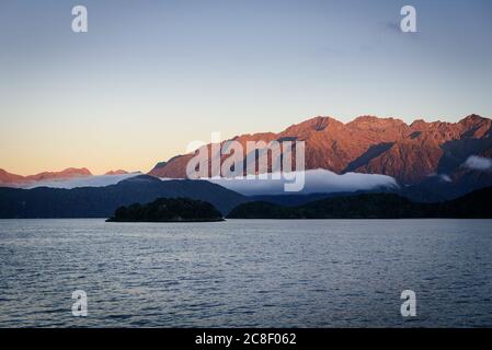 Sonnenaufgang am Lake Manapouri auf der Südinsel Neuseelands. Niedrige Wolken hängen in den rot beleuchteten Bergen auf einer fast vormittags Kreuzfahrt, Stockfoto