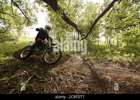 Sportlicher Mann im Helm Motorrad fahren und lehnt sich ab es wiederum beim Überqueren Waldstraße Stockfoto