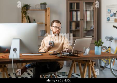 Konzentriertes Design Freiberufler in Brillen sitzen am Holztisch im Home Office und mit Computern beim Kaffee trinken Stockfoto