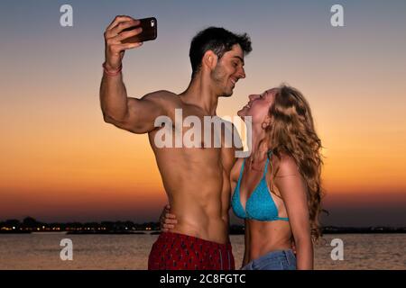 Junges Paar nimmt Selfie im Abendlicht - zwei lächelnde junge Liebhaber machen Selfie am Strand im Sonnenuntergang Stockfoto