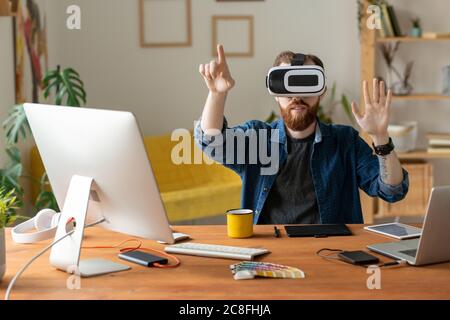 Junger bärtiger Mann berührt unsichtbaren Bildschirm während der Arbeit an virtuellen Design in Virtual Reality-Headset Stockfoto