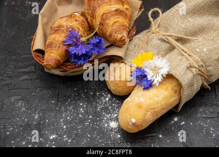 Baguettes in grobes Leinen eingewickelt und ein Korb mit Croissants auf schwarzem Hintergrund. Stockfoto