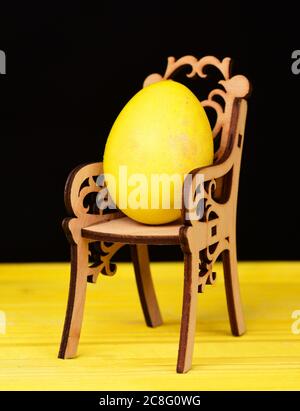 Stuhl mit schönen geschnitzten Mustern aus Holz und hell gelb bemalten Ei für Ostern auf hellen Holztisch isoliert auf schwarzem Hintergrund, close up. Konzept für Osterferien Stockfoto