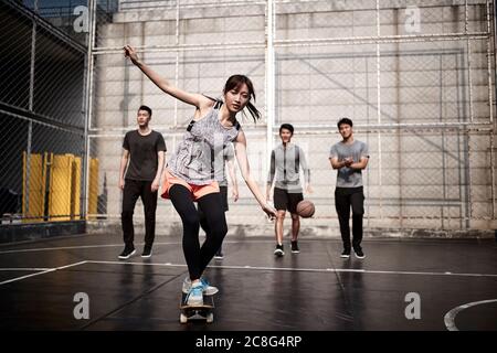 Junge asiatische Frau Skateboarder Skateboarding im Freien mit Freunden beobachten von hinten