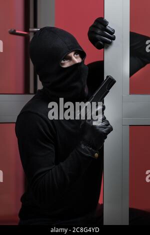 Hausraub durch einen Mann in einer schwarzen Jacke und schwarzer Maske mit schwarzer Waffe. Einbrecher in einer Maske. Dieb in einer Maske versucht, in das Haus anderer Menschen zu brechen Stockfoto