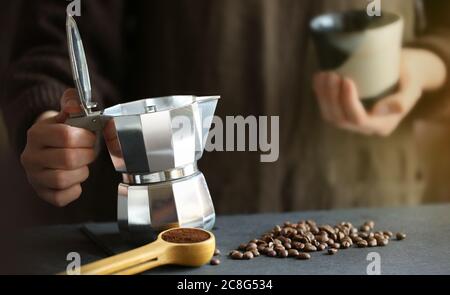 Frau hält in den Händen traditionelle Mokapot Kaffeemaschine bereit, gießen frischen Kaffee in die kleine Tasse. Kaffeebohnen und gemahlener Kaffee auf dem Hintergrund. Stockfoto