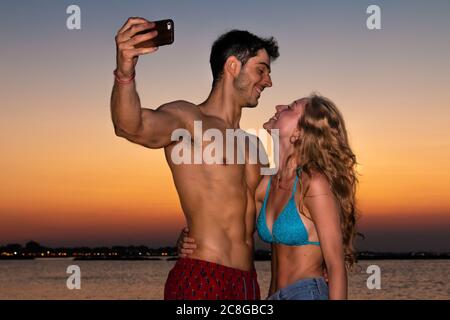 Junges Paar nimmt Selfie im Abendlicht - zwei lächelnde junge Liebhaber machen Selfie am Strand im Sonnenuntergang Stockfoto