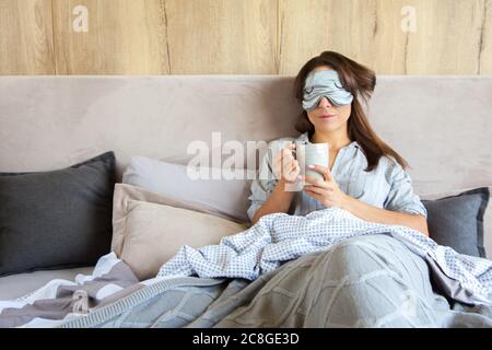 Guten Morgen! Frau wachte im Bett auf. Frau trinkt Kaffee im Bett. Graue Bettwäsche. Augenfleck. Stockfoto