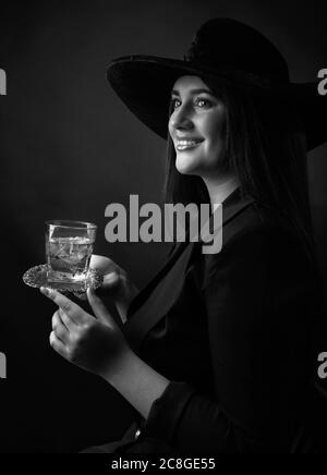 Attraktive Mädchen mit alkoholischen Getränk in gefrorenem Glas. Schöne Brünette in schwarzem Hut hält Glas mit kalten Cocktail. Schwarz und Weiß. Stockfoto