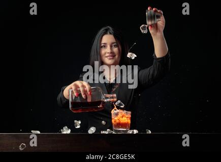 Das attraktive Barkeeper-Mädchen gießt ein alkoholisches Getränk in ein Glas und wirft Eisstücke. Schöne Brünette in schwarz Cocktail vorbereiten. Stockfoto