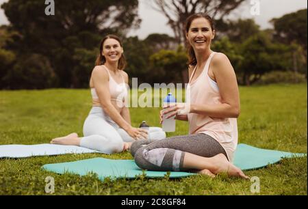 Lächelnde Fitness-Frauen sitzen auf Yogamatten in einem Park Trinkwasser. Zwei fröhliche Freundinnen entspannen nach dem Training. Stockfoto