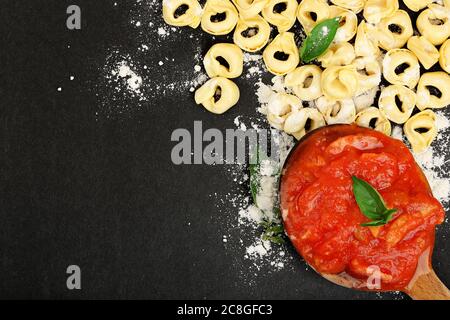 Rohe Tortellini Pasta und Schöpflöffel mit Sauce auf schwarzer Oberfläche Stockfoto