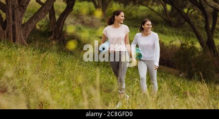 Lächelnde Fitness-Frauen, die mit Yogamatten in einem Park spazieren. Zwei fröhliche Freundinnen auf dem Weg zum Training im Park. Stockfoto