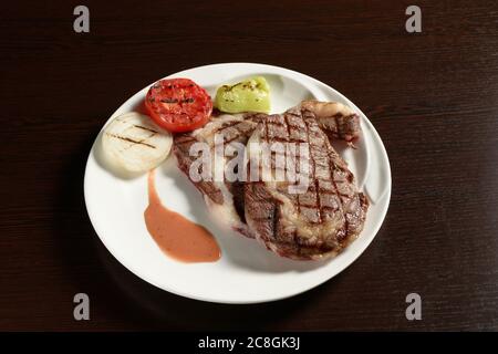 Zwei frittierte Rindersteaks aus Marmor mit gegrilltem Gemüse aus Tomaten, Zwiebeln und Paprika auf einem runden Teller auf einem Holztisch. Fotos für Restaurant und ca. Stockfoto